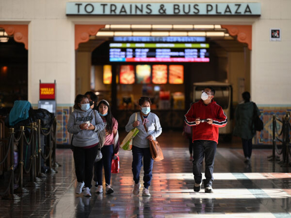Five people walking through a train station, four of them wearing masks