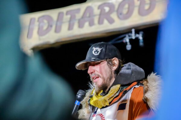 a musher with a microphone under the Iditarod burled arch