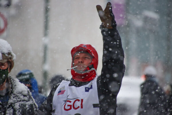 A man wearing a red cap and with a white cigarette in his mouth raises his hand as heavy snow falls