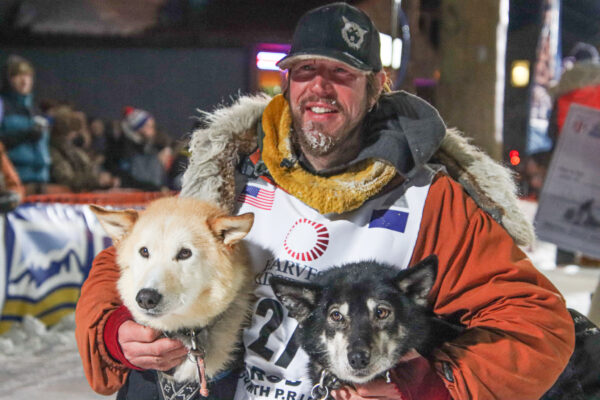 A man in an orange parka holds his dogs on either arm and poses for a photo