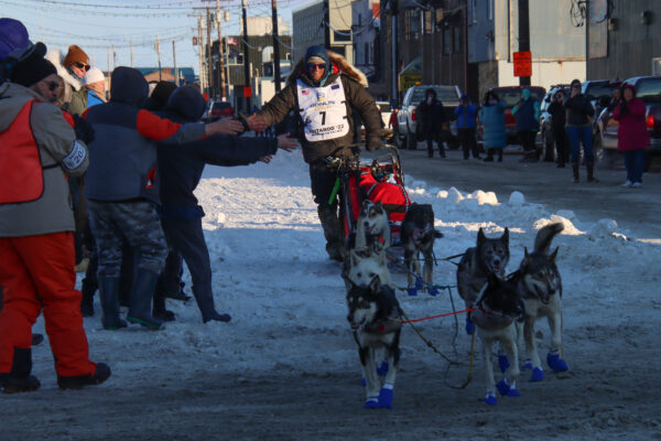 A man high fives a crowd on the back of his sled pulled by a dog team