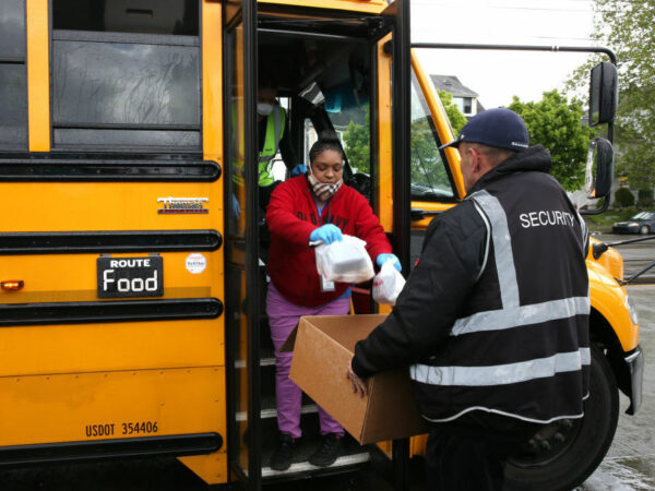 A woman stands on the steps of a school bus and places bagged lunches in a box held by a security guard standing outside