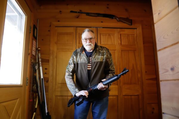 a person wearing camo, jeans, and an American flag shirt holds a firearm near a door and another firearm is visible above him