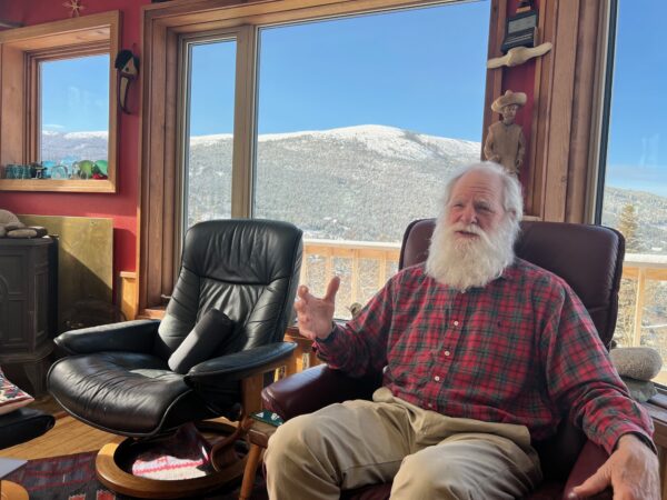 A man with a white beard gestures with his hand in front of a large window and a snowy mountain.