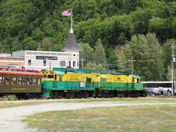 Two green-and-yellow train engines with White Pass markings in front of an old-timey station for tourists