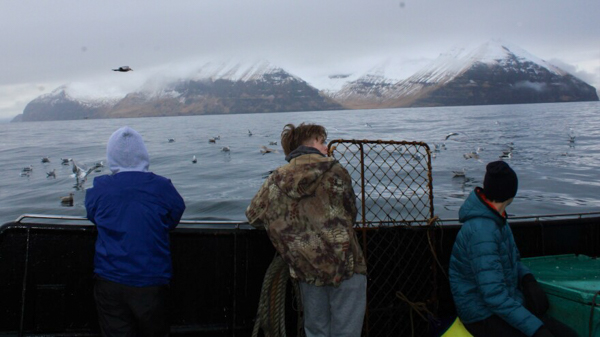 Three teenagers looking off the side of a boat back toward Unalaska