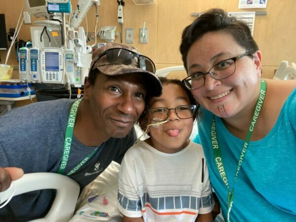A boy sitting up in a hospital bed with his parents on either side of him