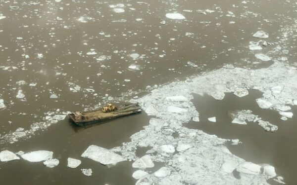 A barge with an excavator on it surrounded by chunks of ice on a river