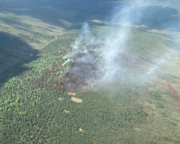 Smoke rises from a fire in trees. Red lines of retardant can be seen along the flanks of the fire. Yellow rain flies mark where firefighters camp’s are locate