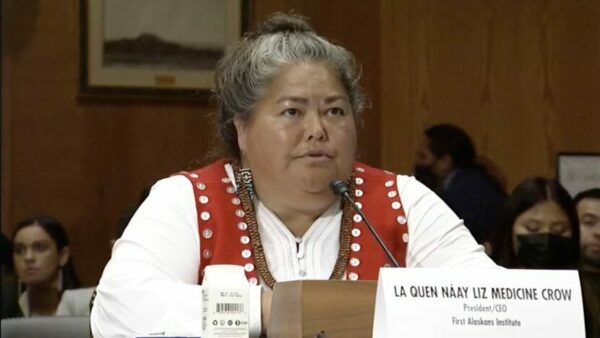 La Quen Náay Liz Medicine Crow sits at a table, speaking into a microphone